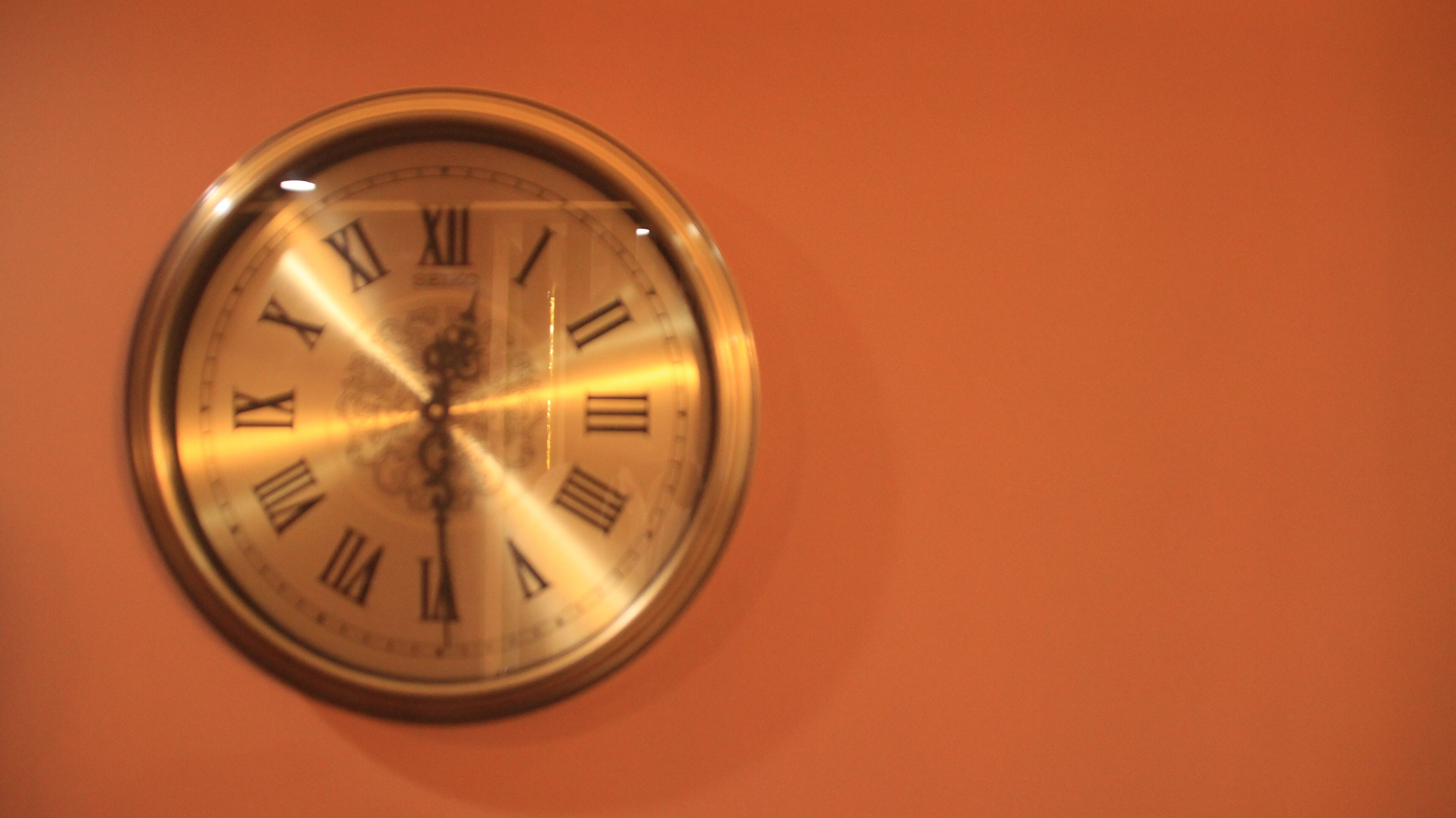 Orologio a parete rotondo con numeri romani e design a lancette centrali.