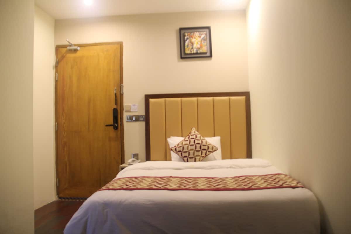 A hotel room with a wooden door, a bed with a patterned pillow, and a framed picture on the wall.