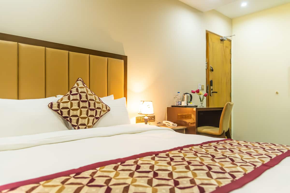 A hotel room with a large bed, a wooden headboard, a desk with a chair, a lamp, and a wooden door.