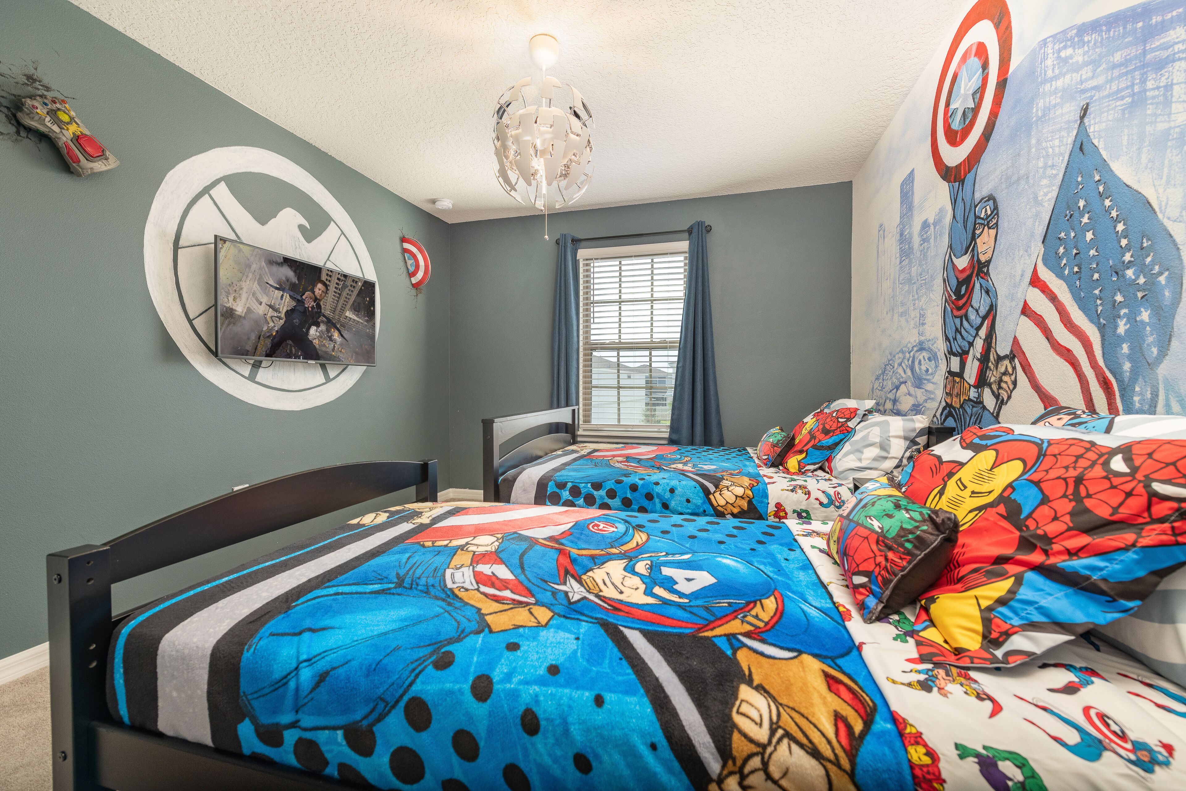 A bedroom with a bed featuring superhero-themed bedding, a circular wall decoration, a window with blinds, and a ceiling fan.
