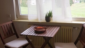 A room with a wooden table and two chairs, a window with white curtains, and a potted plant on the windowsill.
