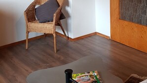 A room with a wooden ceiling, a wicker chair with a cushion, a small table with a magazine, and a bed with a patterned blanket.