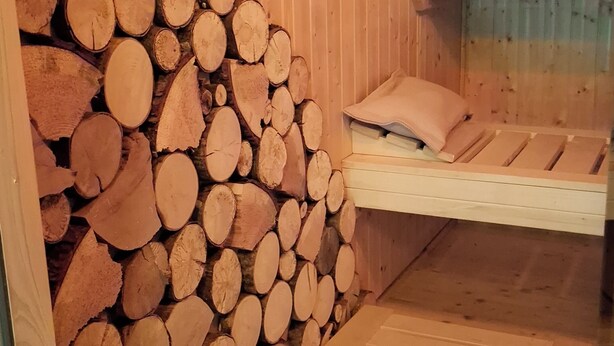 A wooden cabin interior with a bed and a stack of firewood against the wall.