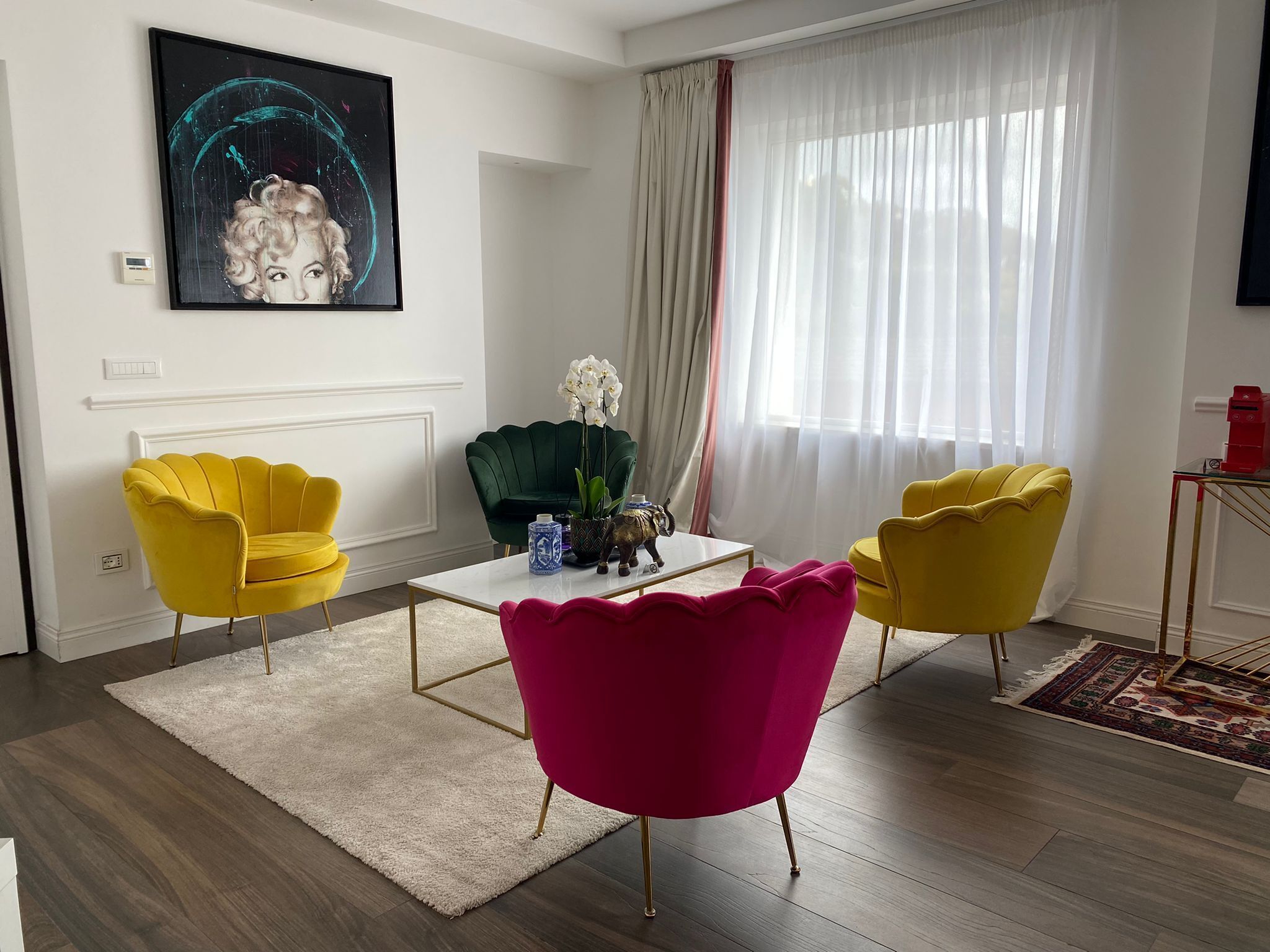 En moderne stue med en hvid sofa, en gul lænestol, en lyserød lænestol, en grøn lænestol og et sofabord. Der er et billede på væggen og et vindue med lette gardiner.