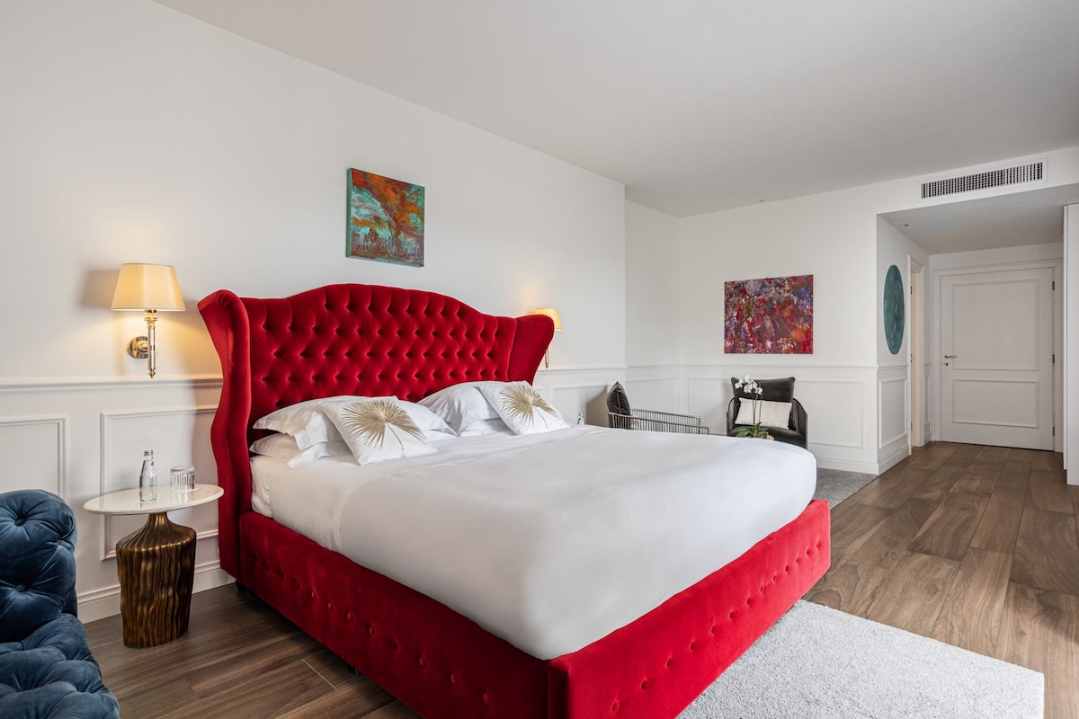 A spacious bedroom with a large red upholstered bed, white bedding, and a small round table with a lamp.
