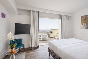 A hotel room with a large bed, a TV, a desk with a chair, a view of the city, and a balcony with outdoor seating.