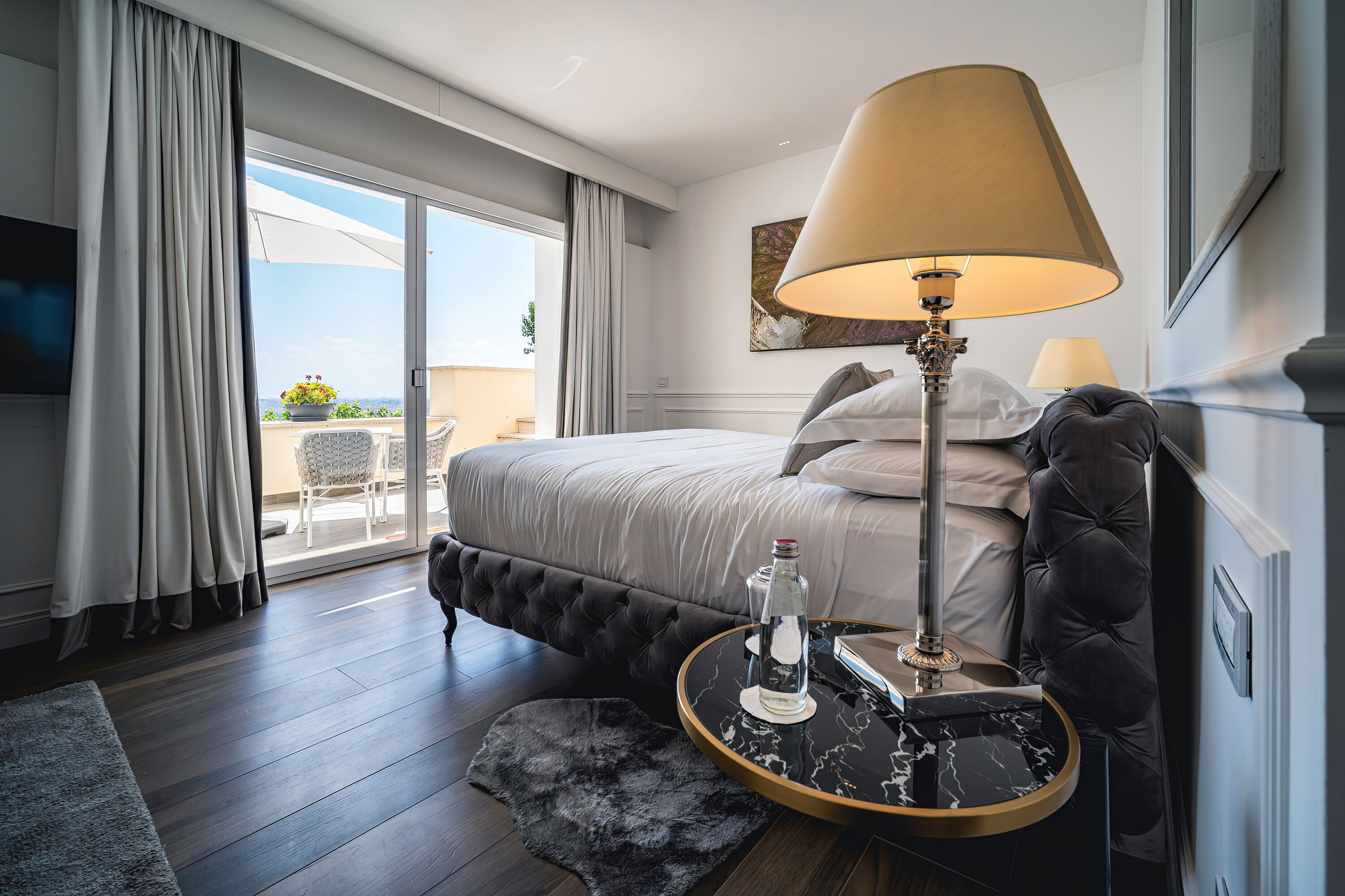 A modern hotel room with a large bed, a round side table with a lamp, a glass bottle, and a view of the outdoors through a sliding glass door.