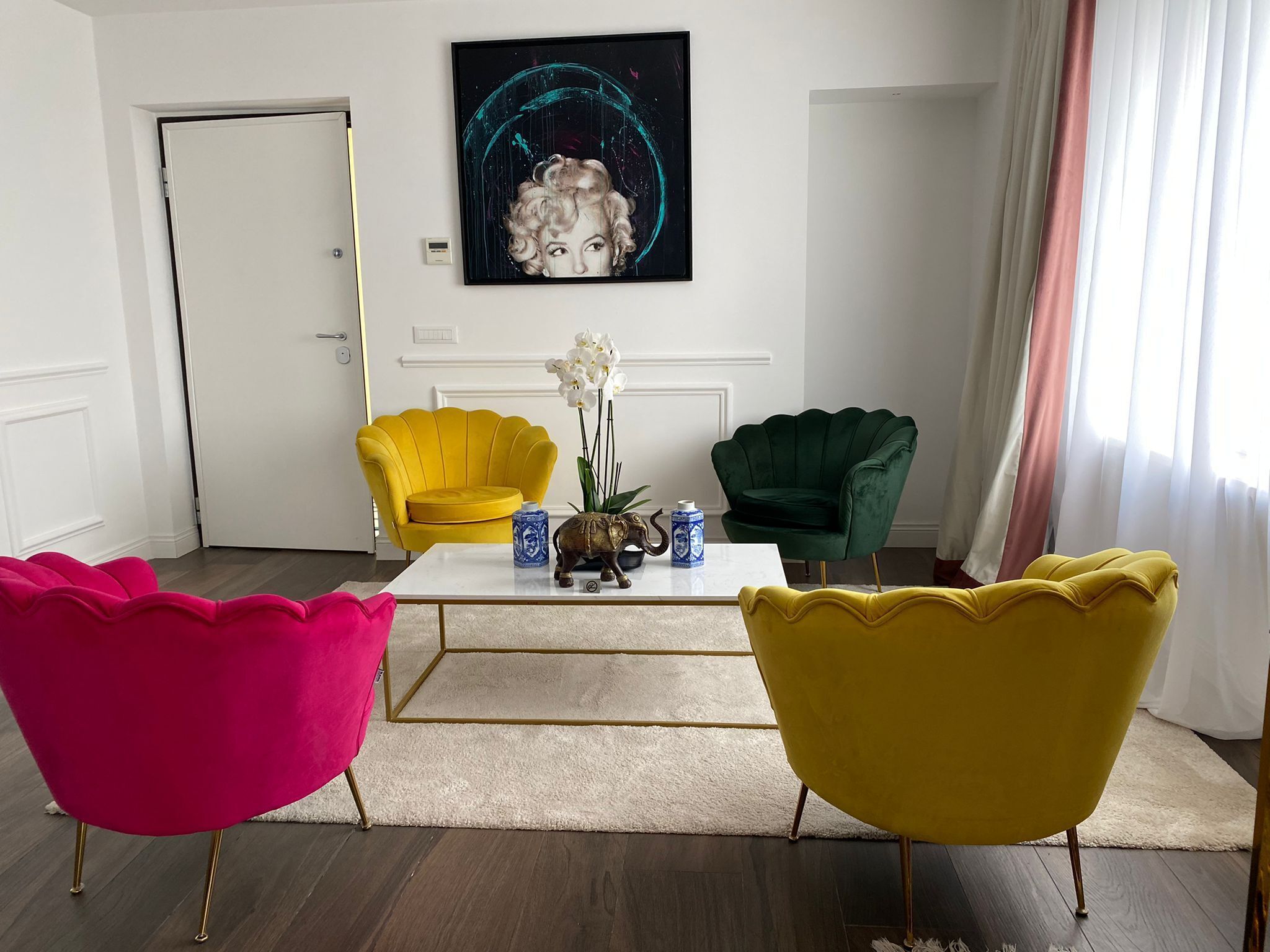 A modern living room with a white coffee table, two vibrant armchairs, a green armchair, and a painting on the wall.