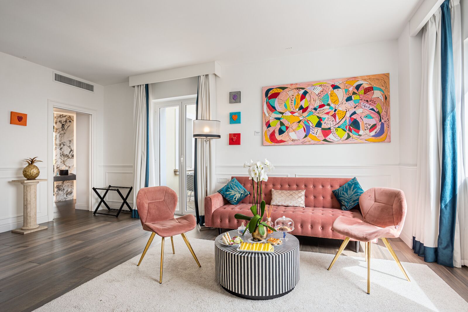 A modern living room with a pink sofa, two pink chairs, a striped ottoman, and a colorful abstract painting.