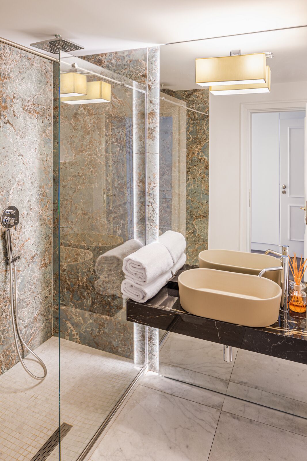Une salle de bain moderne dotée d’une douche à parois de verre, de murs en marbre, d’un grand miroir et d’un plan de toilette noir avec un lavabo beige.