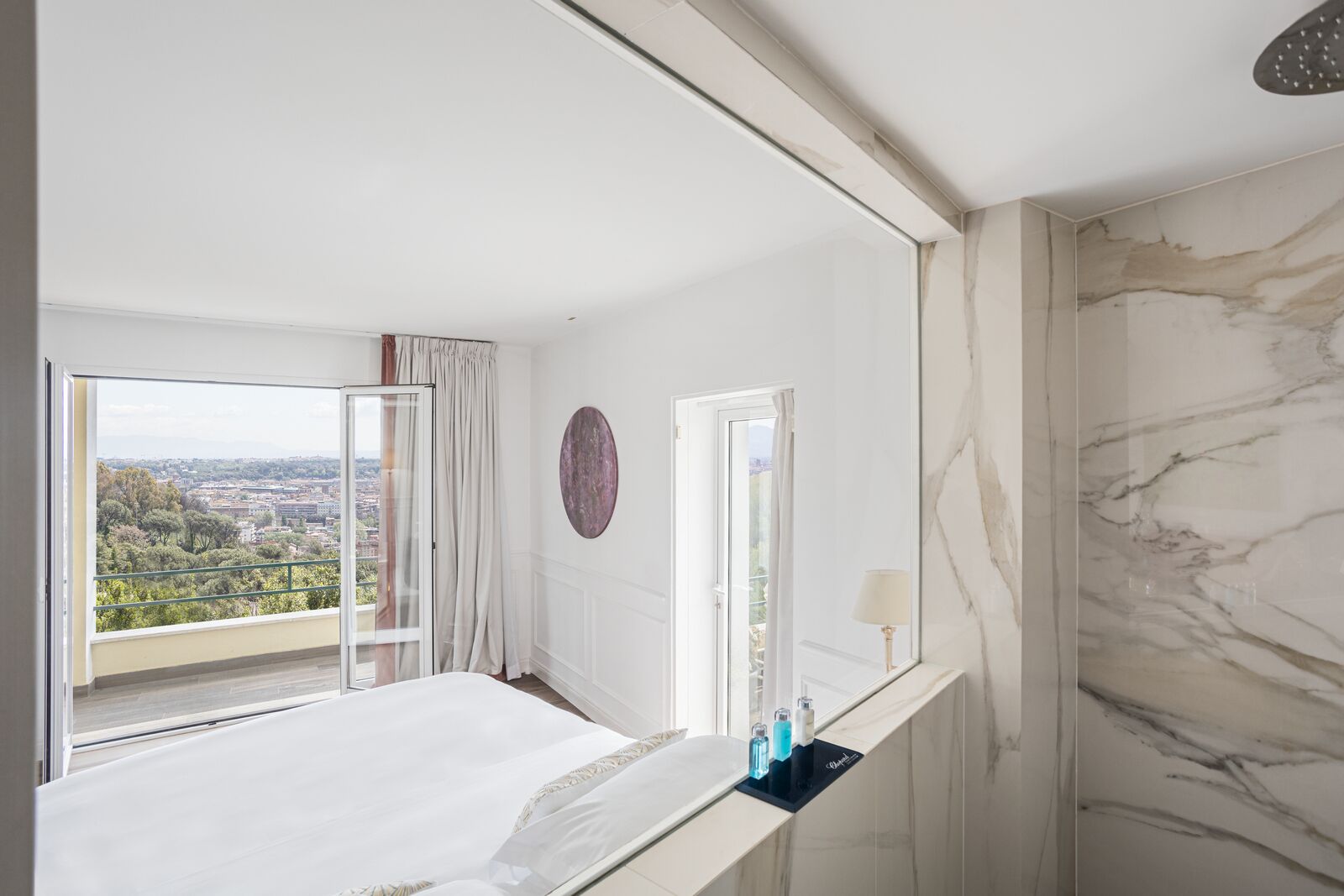 A bedroom with a bed, a bathroom with a marble wall, and a view of a cityscape through a window.