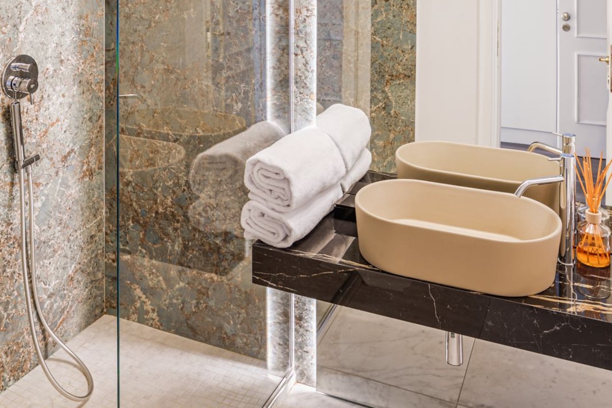 Un baño moderno con ducha de vidrio, paredes de mármol, un espejo grande y encimera negra con lavabo color beige.