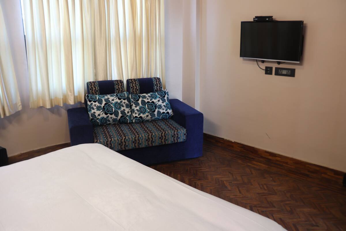 A hotel room with a bed, a sofa with patterned cushions, a television on the wall, and a wooden floor.