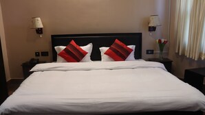 A neatly made bed with white linens and two red and white pillows. There are two bedside tables with lamps and a vase of flowers on one of them. - Rinchen Hotel (Kathmandu)