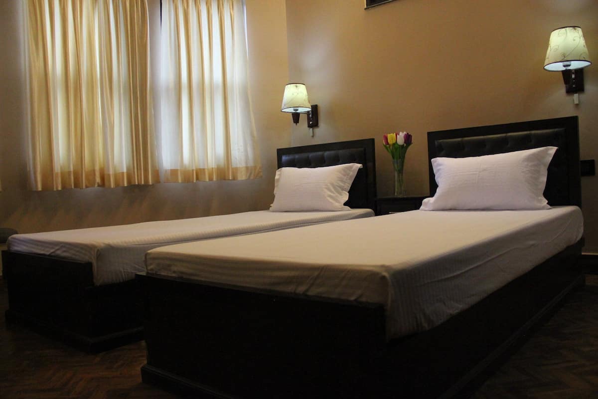 A hotel room with two single beds, each with a white pillow and a small bedside table. There are two wall-mounted lamps on either side of the beds.