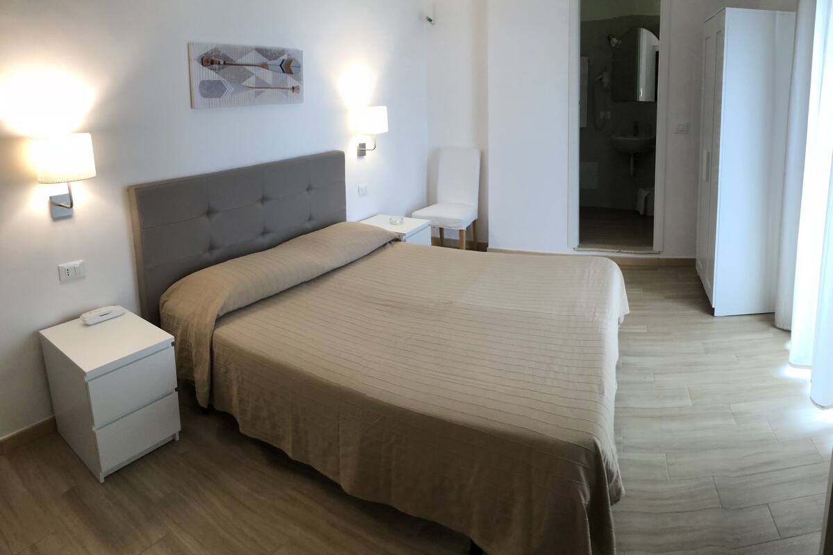 Een hotelkamer met een groot bed, nachtkastjes, een wandlamp en een badkamer zichtbaar door een open deur.