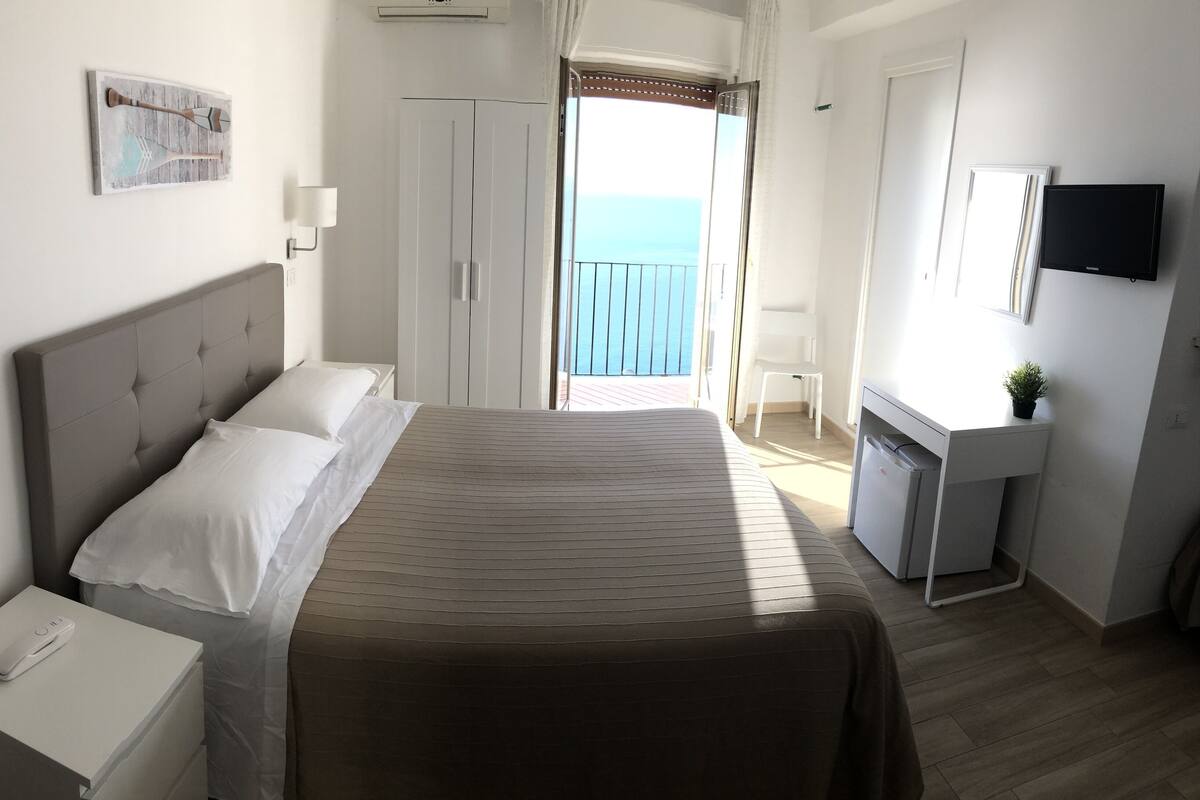 Een hotelkamer met een bed, nachtkastje, lamp, televisie en uitzicht op zee door een open deur.
