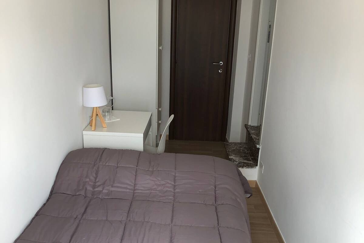 Een slaapkamer met een groot bed, een nachtkastje met lamp, een kledingkast en een deur op de achtergrond.