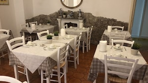 A dining area with white tables and chairs, each set with plates, cups, and cutlery. There are potted plants on the tables, and a stone wall with a fireplace in the background. - Villa Jole Taormina (Taormina)