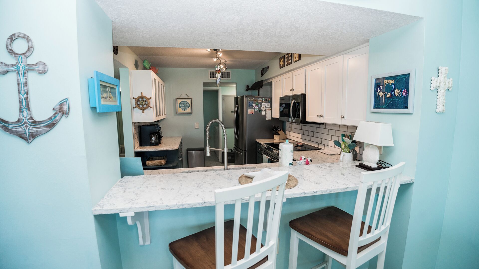 A compact kitchen with white cabinets, a marble countertop, and a sink. There are two white chairs at the counter.