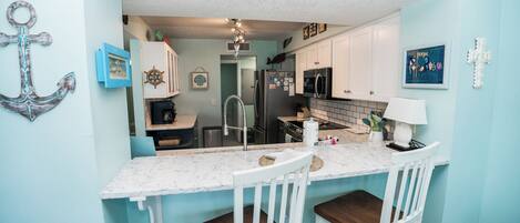 A compact kitchen with white cabinets, a marble countertop, and a sink. There are two white chairs at the counter.