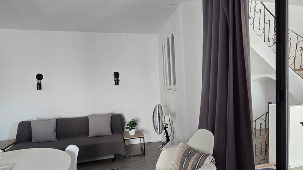 A modern living room with a grey sofa, a white dining table, and a white armchair. There is a small side table with a plant, a standing fan, and a large window with curtains. - Apartment 43m2 sea view (Altea)