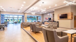 A modern lounge area with sofas and tables, large windows, and a mounted TV.