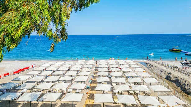 A beach with rows of white umbrellas and sun loungers, a clear blue sea, and a rocky shoreline.