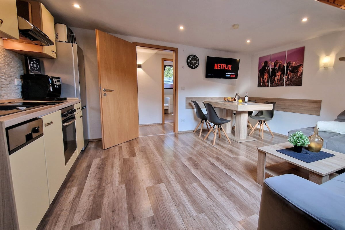 A modern kitchen with a microwave, stove, and oven. There is a dining area with a table and chairs, and a living area with a sofa and a coffee table. A TV is mounted on the wall, and there are decorative items on shelves.