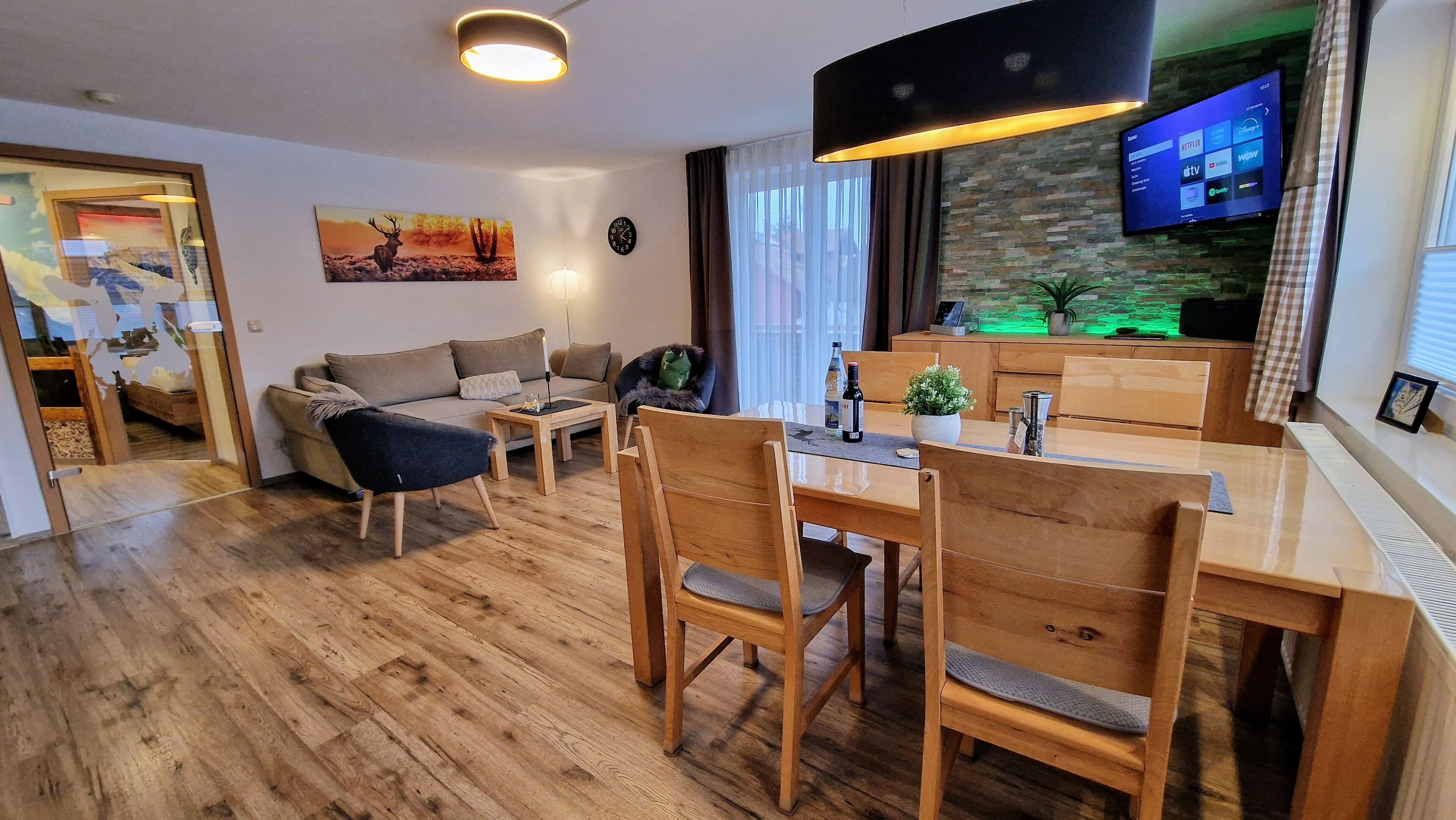 Una sala de estar moderna con un sofá, una mesa de comedor con sillas, un televisor montado en la pared y un reloj.