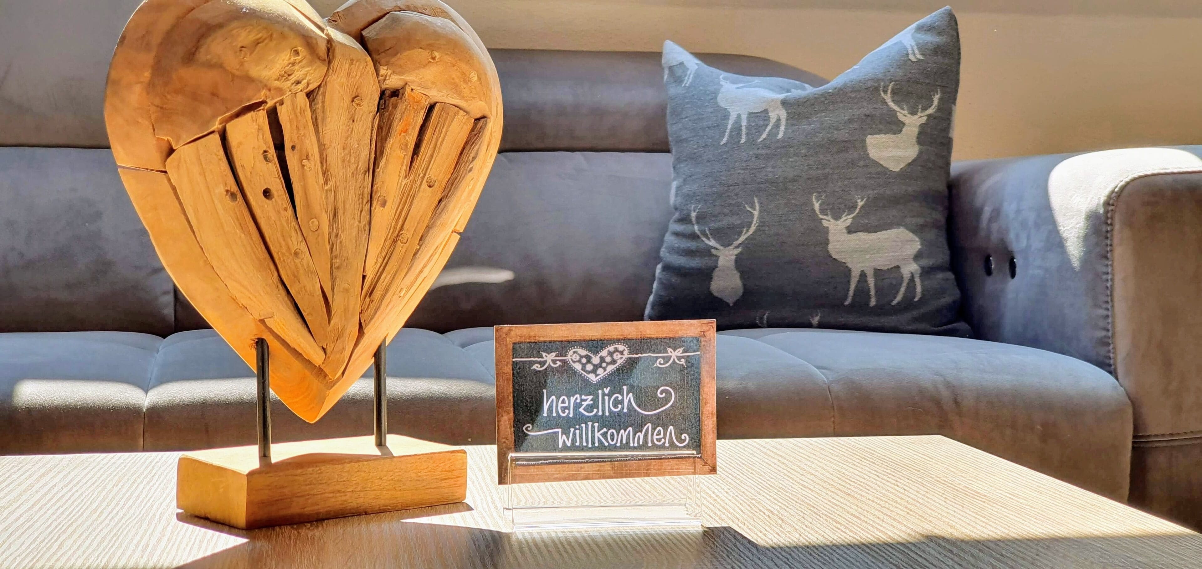 Una escultura de madera con forma de corazón sobre una base de madera, un cartel con la inscripción "herzlich willkommen" (bienvenidos) y un sofá con un cojín que muestra siluetas de ciervos.