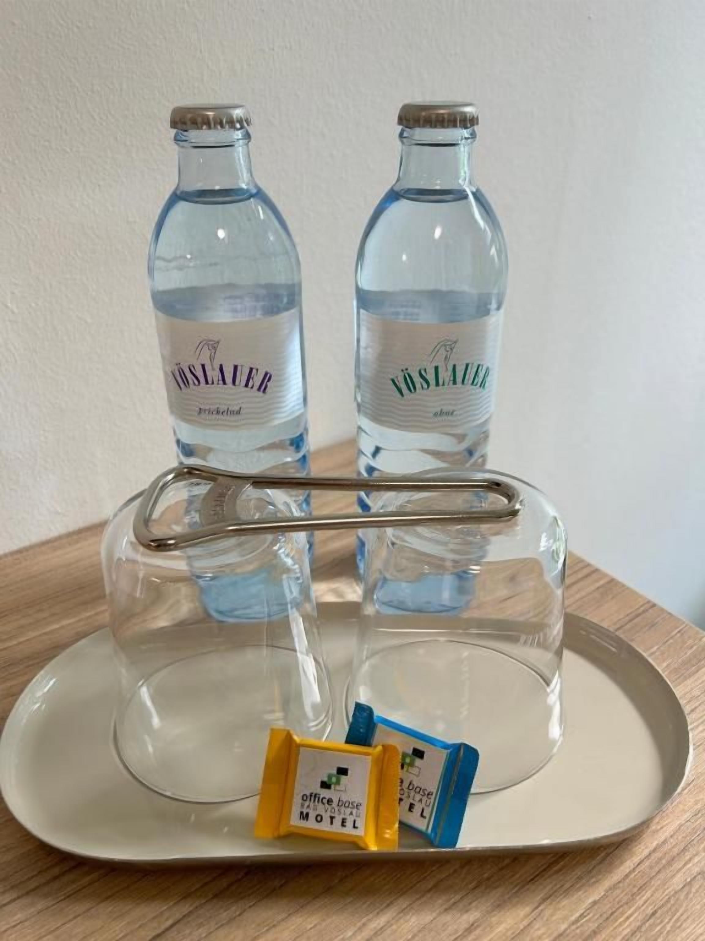 Two clear water bottles labeled 'VÖSLAUER' on a tray with a metal stand. Below the bottles, there are two small packets, one yellow and one blue, with text on them.