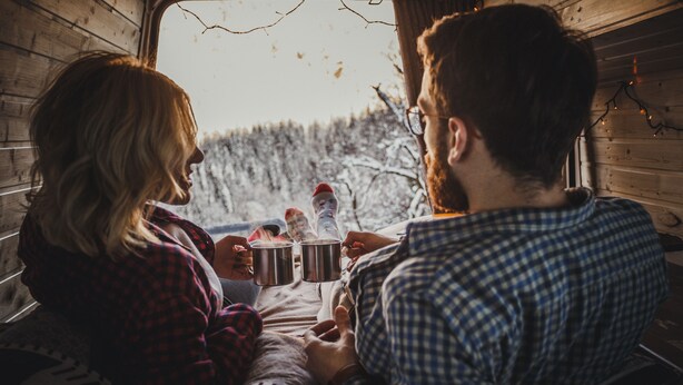 A cozy cabin interior with wooden walls, a window overlooking a snowy forest, and two people holding mugs. - Self-built campervan (Kutaisi)