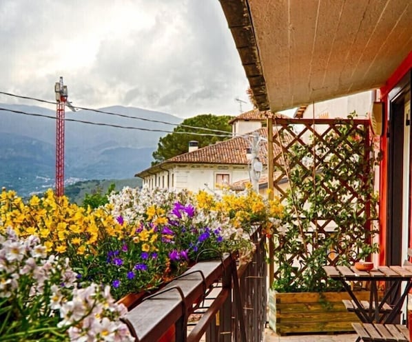 A balcony with a wooden railing overlooking a garden with various flowers, a building with a tiled roof, and a red telephone pole in the background. - Il Piccolo Tibet (L'Aquila)
