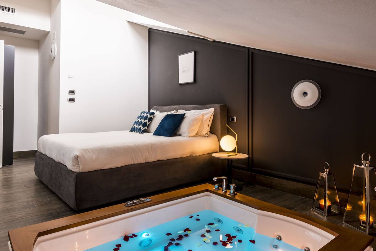 Ein modernes Hotelzimmer mit einem großen Bett, einer Badewanne mit Rosenblüten und einem Nachttisch mit Lampe.