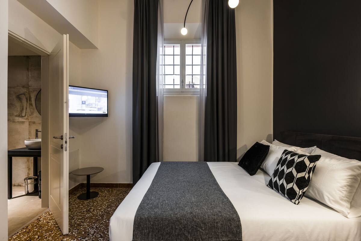 Ein Hotelzimmer mit Bett, Fernseher, Fenster mit Vorhängen und einem kleinen Tisch.