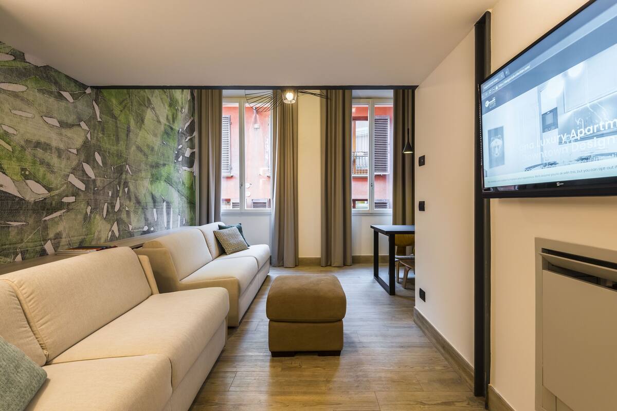 Ein modernes Wohnzimmer mit einem beigefarbenen Sofa, einem kleinen Hocker, einem flachbildfernseher an der Wand und einem großen, abstrakten Gemälde links an der Wand.