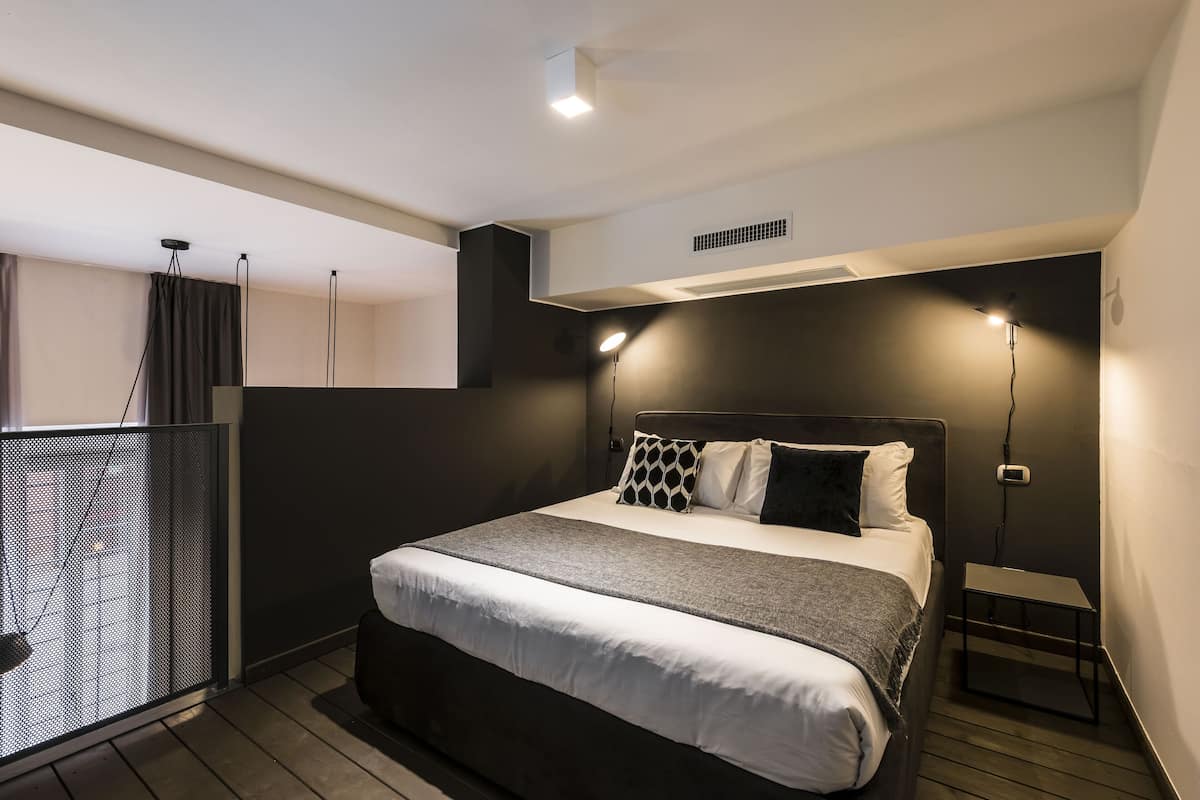 Ein modernes Schlafzimmer mit einem großen Bett, einem kleinen Nachttisch und Blick auf die Stadt durch ein Fenster.