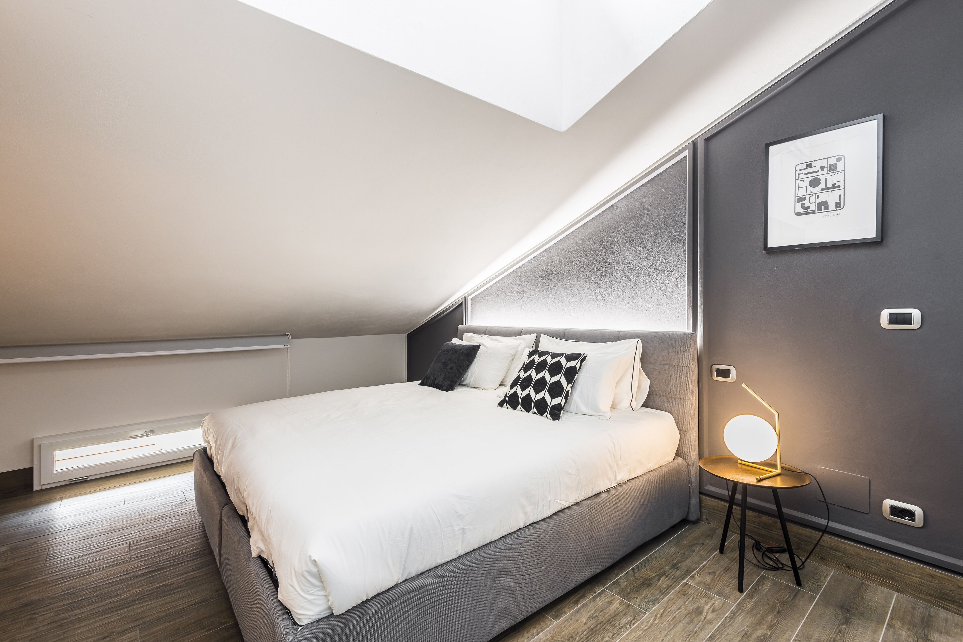 A modern bedroom with a skylight, a bed with white bedding, a small bedside table with a lamp, and a framed picture on the wall.