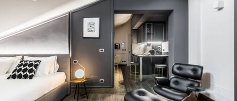 A modern hotel room with a bed, a seating area with a chair, a desk, and a kitchenette.