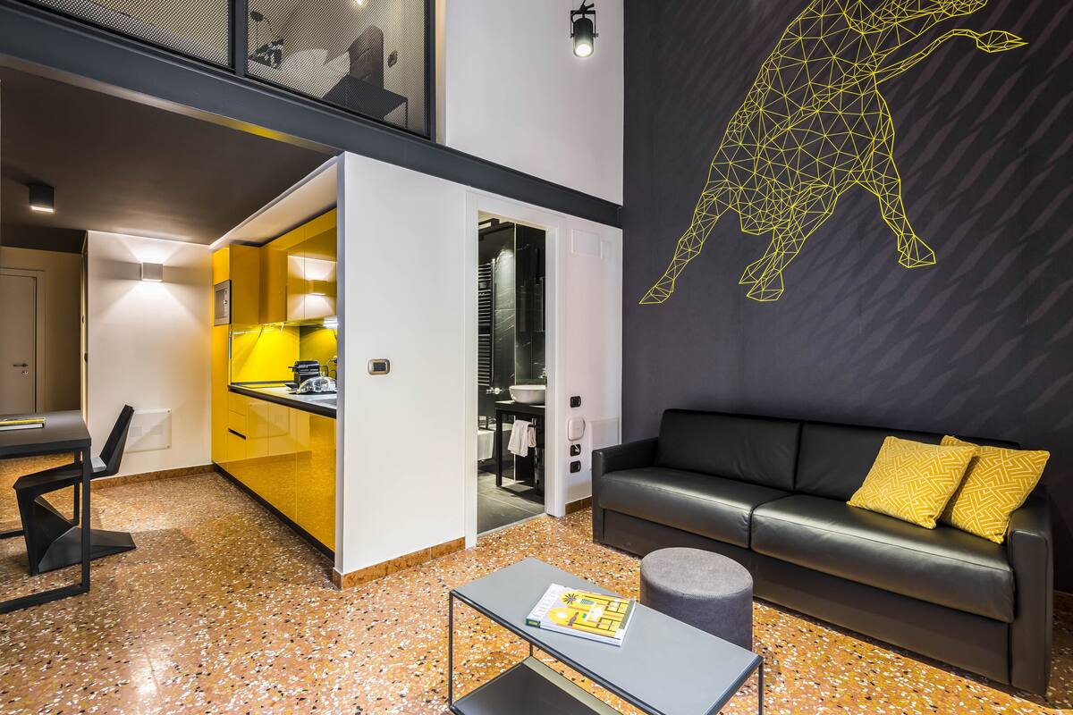Ein moderner Wohnraum mit Küchenbereich, einer Couch mit gelben Kissen und einem Wandbild eines geometrischen Tieres.