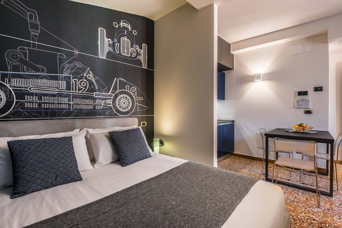 Ein Hotelzimmer mit Bett, Kissen, Wandbild alter Autos, Schreibtisch mit Stuhl und einem kleinen Tisch mit Blumenvase.
