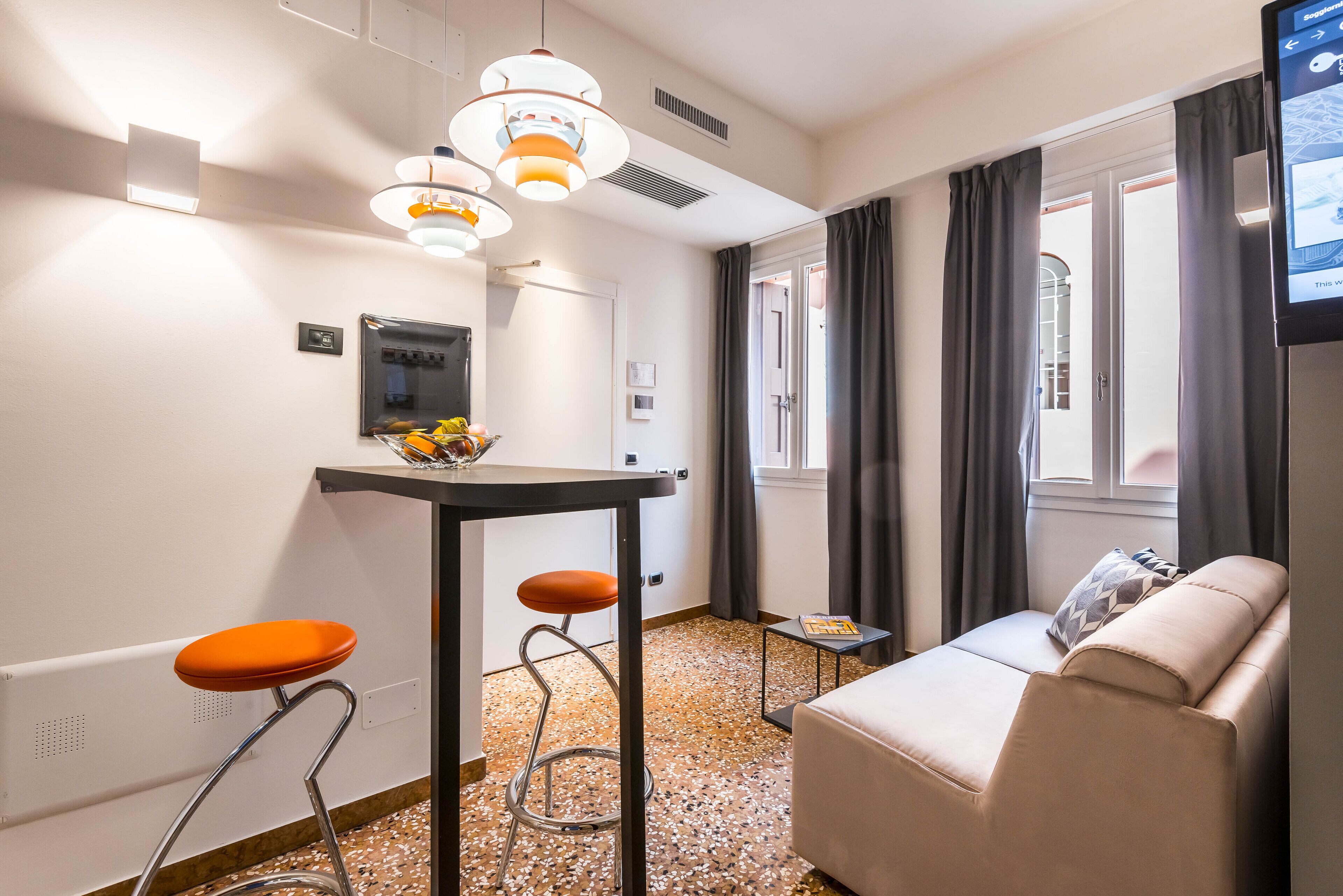 Ein modernes Wohnzimmer mit einer Couch, einem kleinen Tisch und einem Barhocker. Es gibt ein Fenster mit Vorhängen, einen an der Wand montierten Fernseher und eine Deckenleuchte.