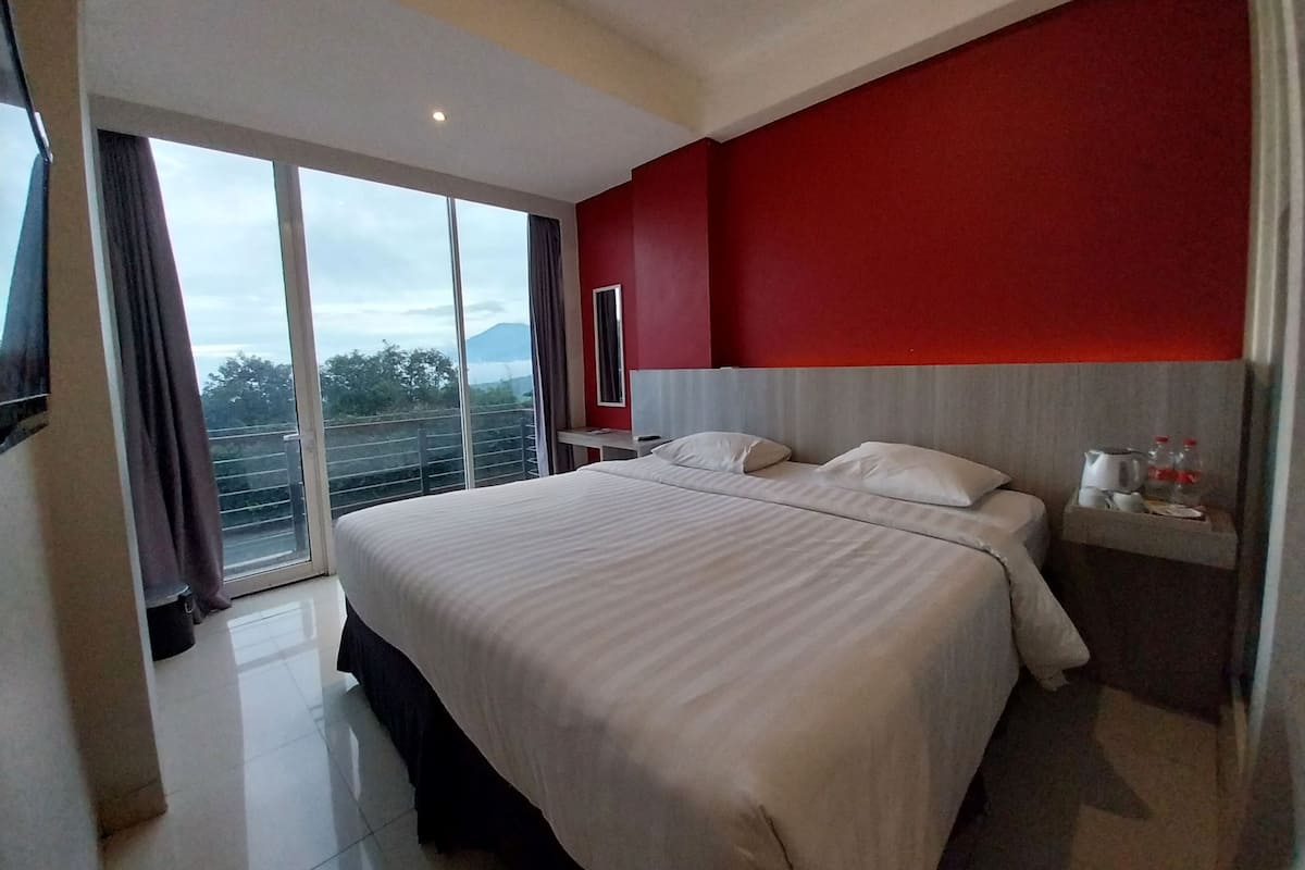 Habitación de hotel con una cama grande, una pared de acento roja, una mesita de noche con hervidor y botellas, y vistas al exterior.