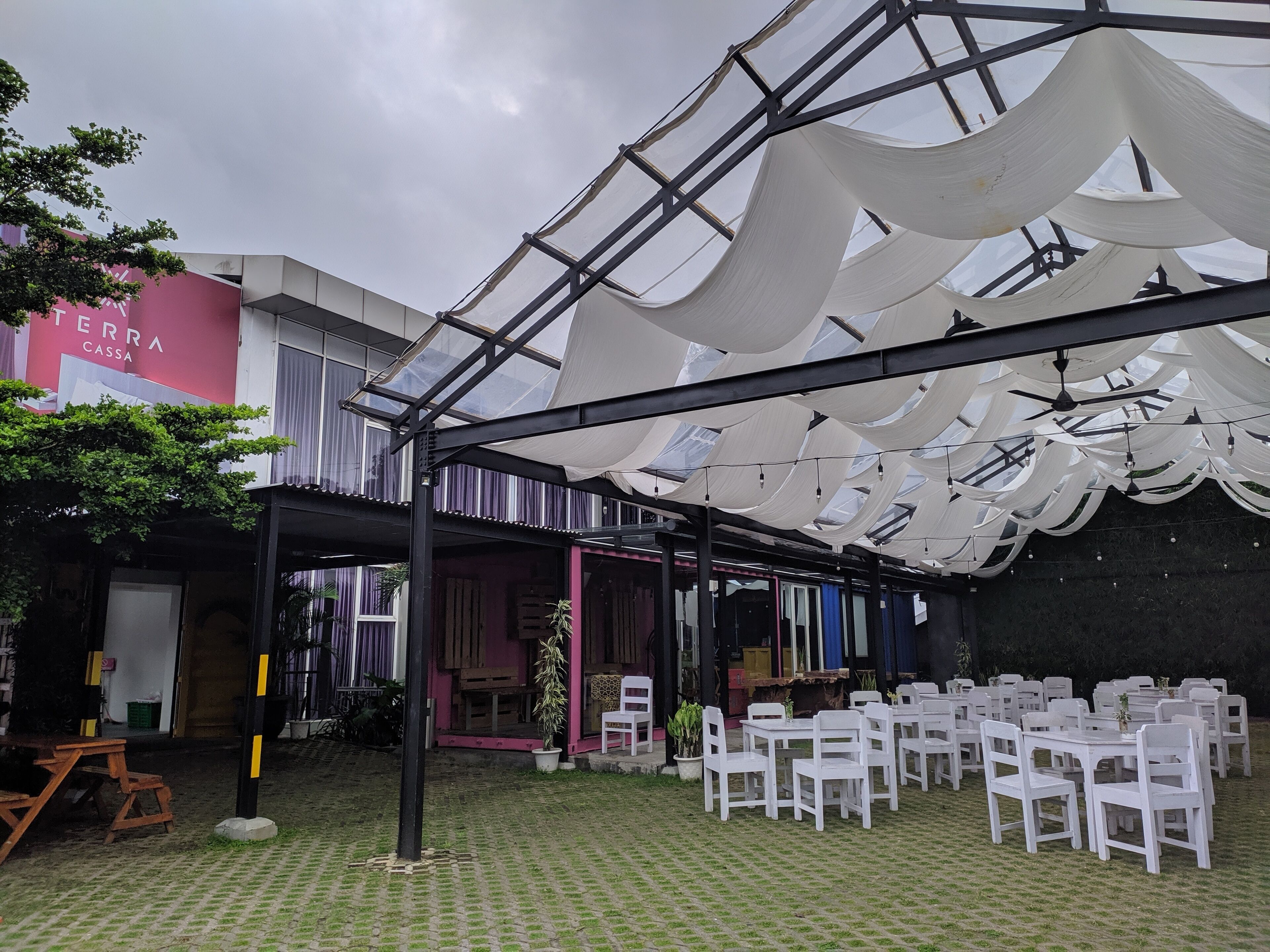 An outdoor patio with white chairs and tables, a covered area with a glass ceiling, and a building with a pink facade in the background.