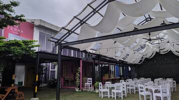 An outdoor patio with white chairs and tables, a covered area with a glass ceiling, and a building with a pink facade in the background.