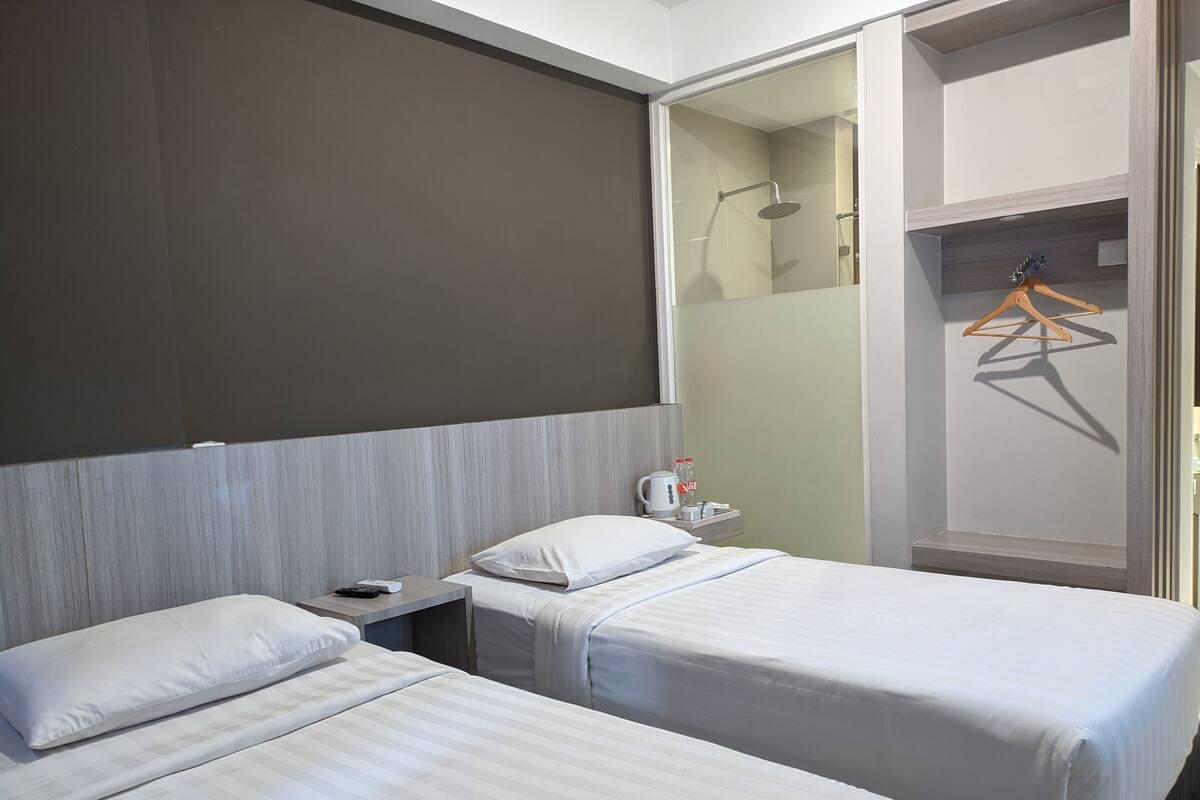 Habitación de hotel con dos camas, una mesita de noche, un espejo, zona de ducha y un perchero de madera.