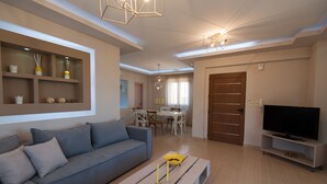 Una sala de estar moderna con un sofá gris, una mesa de centro de madera, un televisor sobre un mueble bajo, y un comedor al fondo.