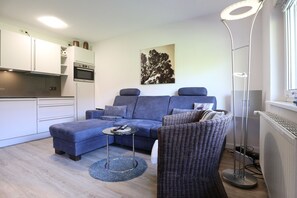 Un espacio habitable pequeño con sofá, una mesita, una silla de mimbre, una lámpara de pie y una zona de cocina.