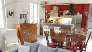 A modern living room with a grey sofa, wooden dining table, and chairs. A kitchen with red accents and a stainless steel refrigerator is visible in the background.